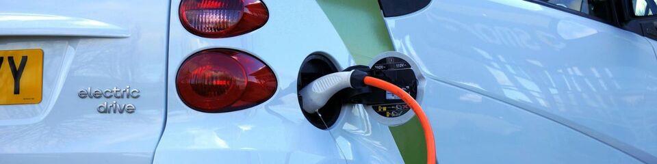 Macchine elettriche_websim1 Auto elettrica collegata a una stazione di ricarica, con il cavo connesso e una luce che indica il progresso della carica.