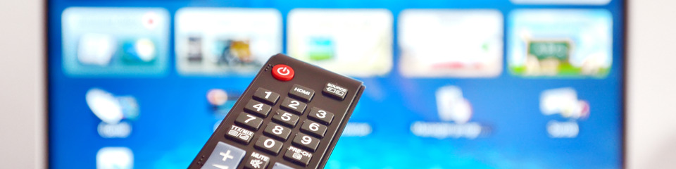 Telecomando in primo piano con pulsanti visibili; sullo sfondo si intravede una TV accesa sfocata.