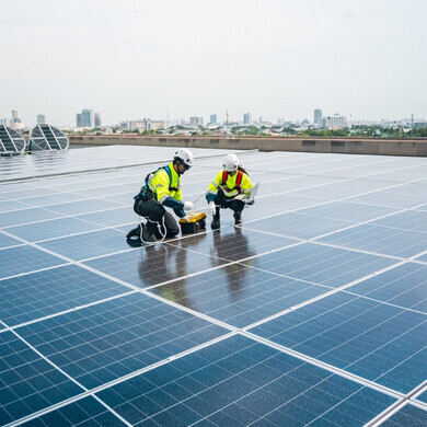 Un uomo e una donna, entrambi con giubbotti fluorescenti e caschi di sicurezza, installano pannelli solari in una giornata soleggiata utilizzando laptop e voltmetro.