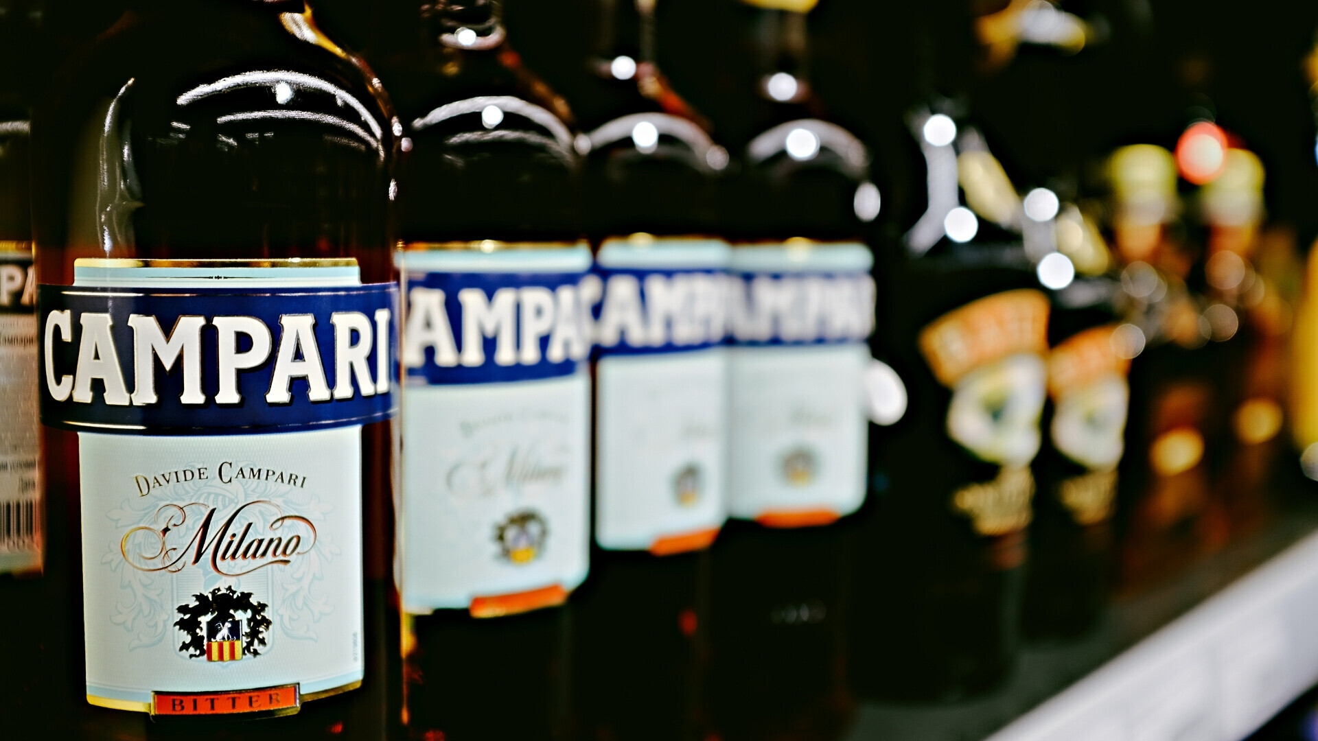 Scena d’insieme con diverse bottiglie Campari, riflessi di luce che valorizzano il design iconico e la trasparenza del vetro.