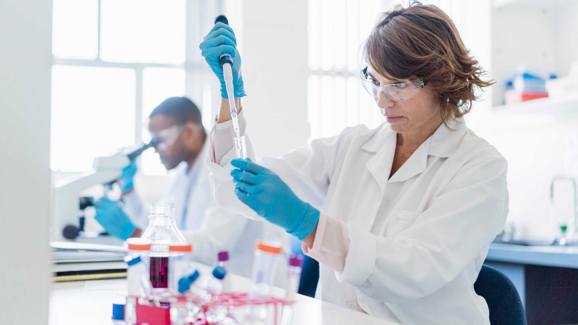 Foto di chimico concentrato che lavora su sostanze chimiche. Scienziata donna utilizza una pipetta con collega sullo sfondo, all’interno di un laboratorio.