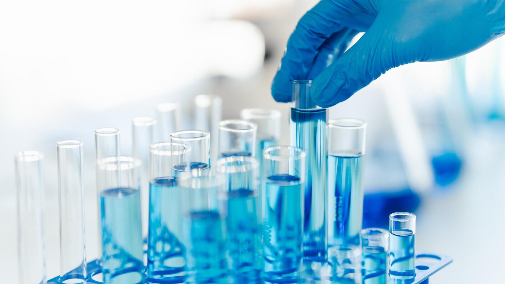 Primo piano della mano guantata di un tecnico di laboratorio che tiene una provetta con liquido blu tra altre provette su uno scaffale, simbolo di ricerca scientifica e analisi chimica in un laboratorio moderno.
