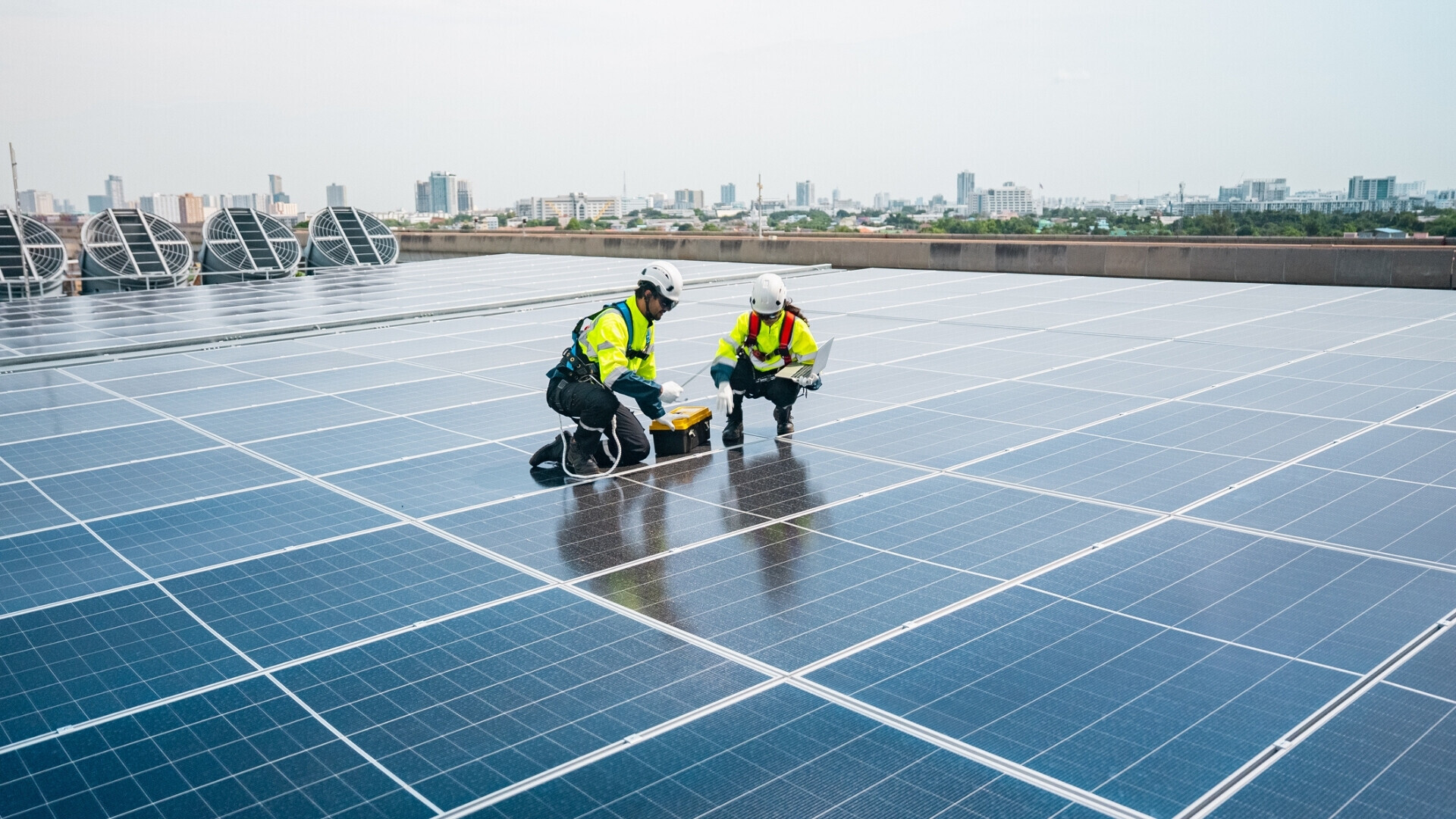 Un uomo e una donna, entrambi con giubbotti fluorescenti e caschi di sicurezza, installano pannelli solari in una giornata soleggiata utilizzando laptop e voltmetro.