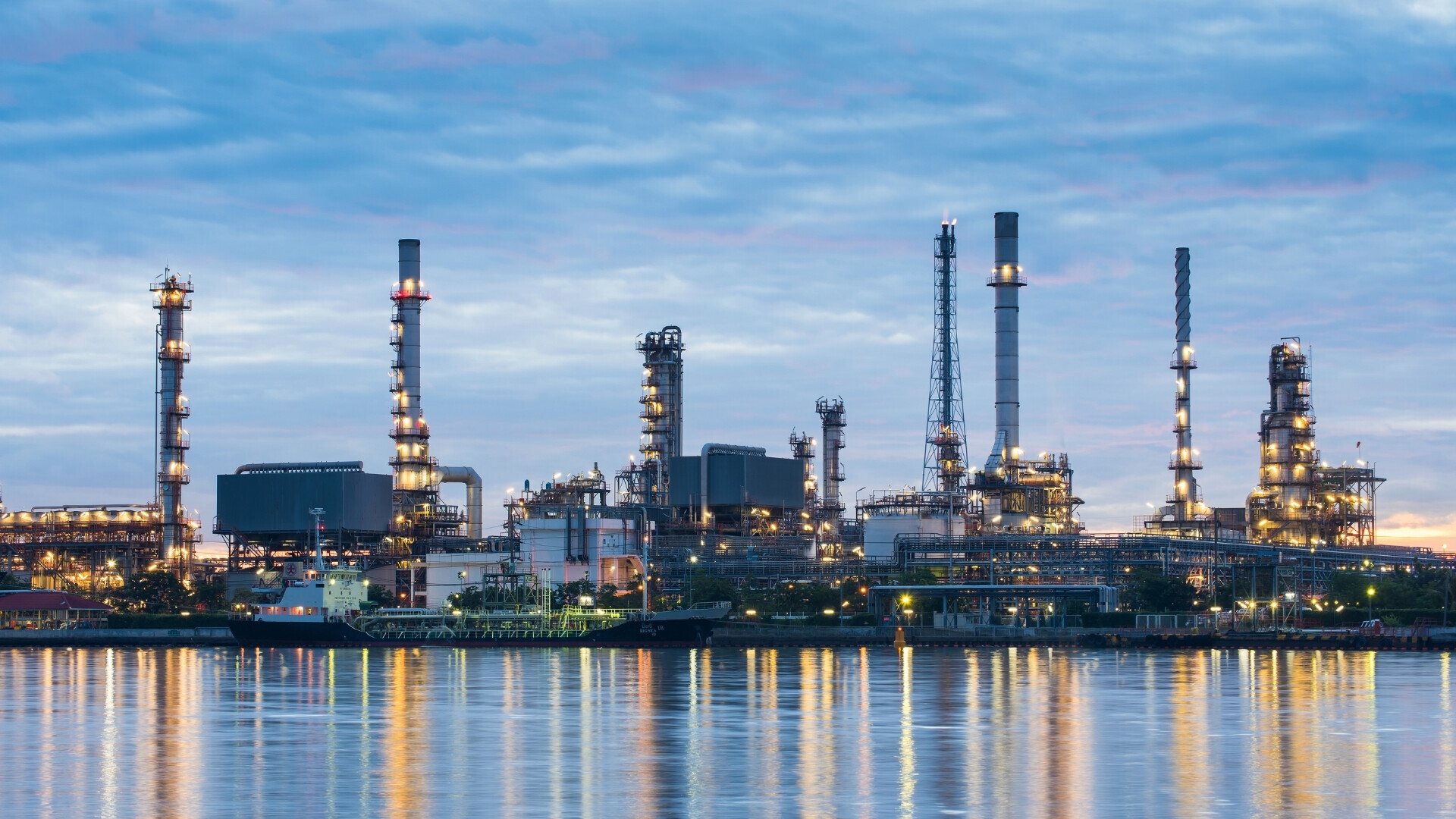 Vista panoramica di una raffineria di gas con torri e impianti illuminati, riflessi sull'acqua calma di fronte, sotto un cielo serale nuvoloso.