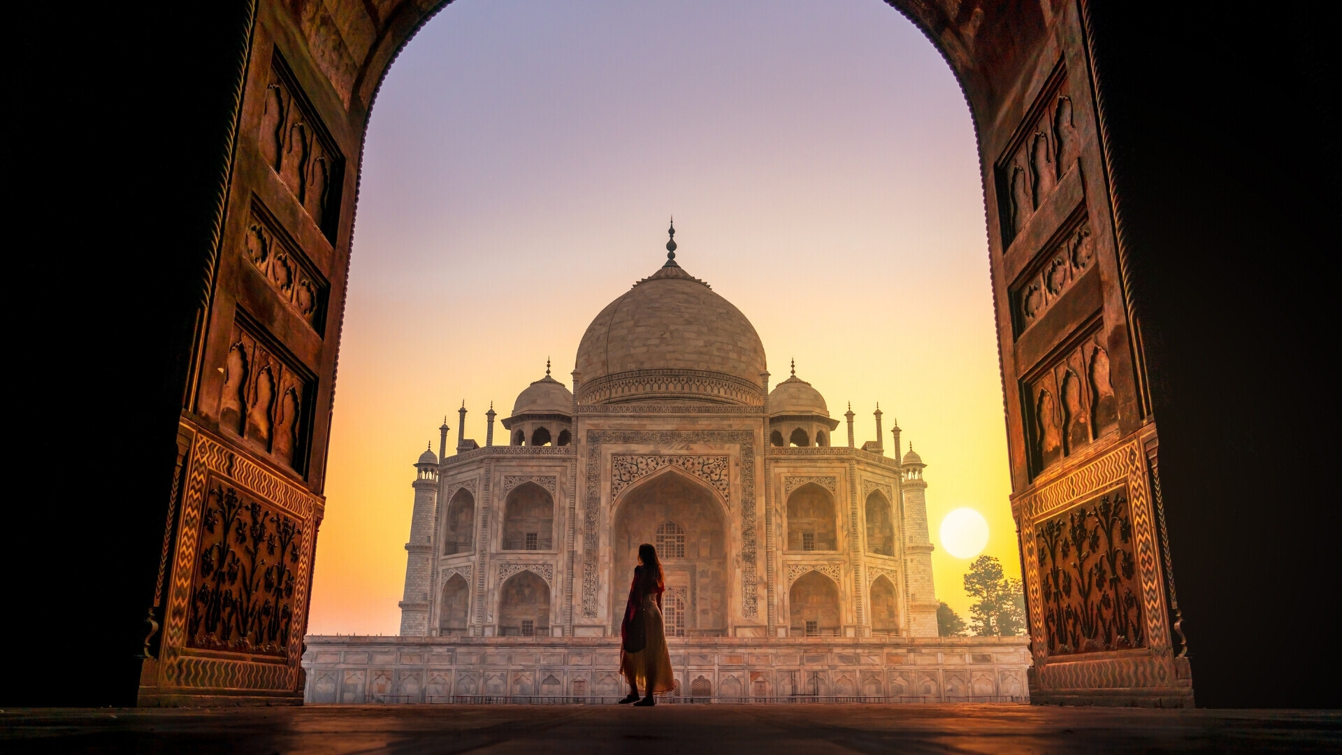 Il Taj Mahal è inquadrato attraverso un grande arco decorato, illuminato dalla luce calda del tramonto. Al centro, in primo piano, si vede la sagoma di una persona che osserva il monumento, mentre il cielo sfuma dal rosa all’oro e il sole basso sull’orizz