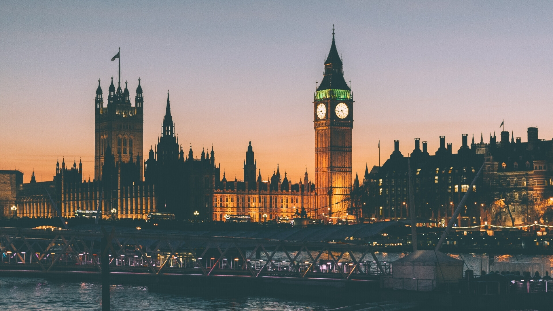 Vista panoramica del Palazzo di Westminster e del Big Ben illuminati al tramonto, con il Tamigi in primo piano e il cielo dai toni caldi che avvolge lo skyline di Londra.