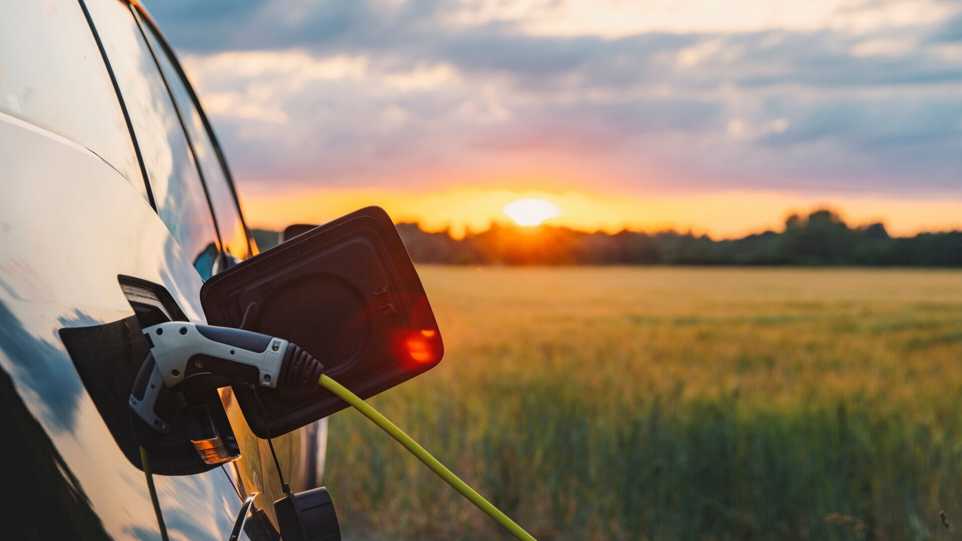 Auto elettrica collegata alla ricarica in campagna al tramonto Auto elettrica in ricarica in un’area rurale al tramonto, con campi e cielo arancione sullo sfondo, rappresentazione di mobilità sostenibile.