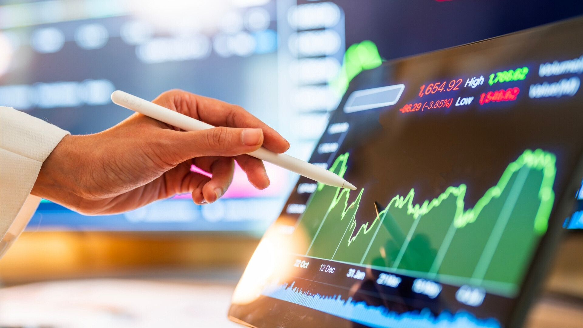 Primo piano di una mano che utilizza una penna digitale per analizzare tendenze finanziarie su un tablet, con grafici di borsa fluttuanti.